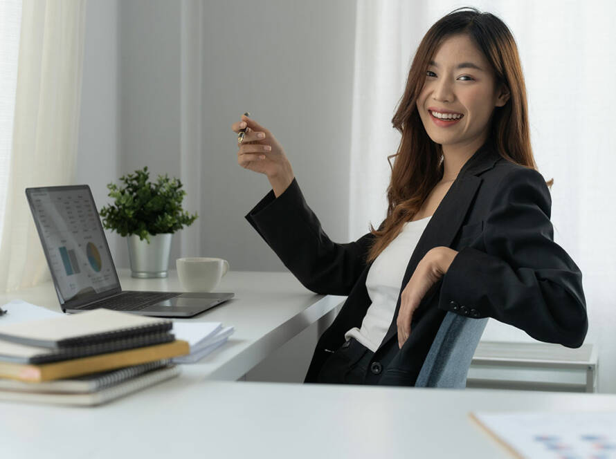 Asian lady smiling while leaning back in her office chair holding a pen, at a desk in front of a laptop with books and paperwork.